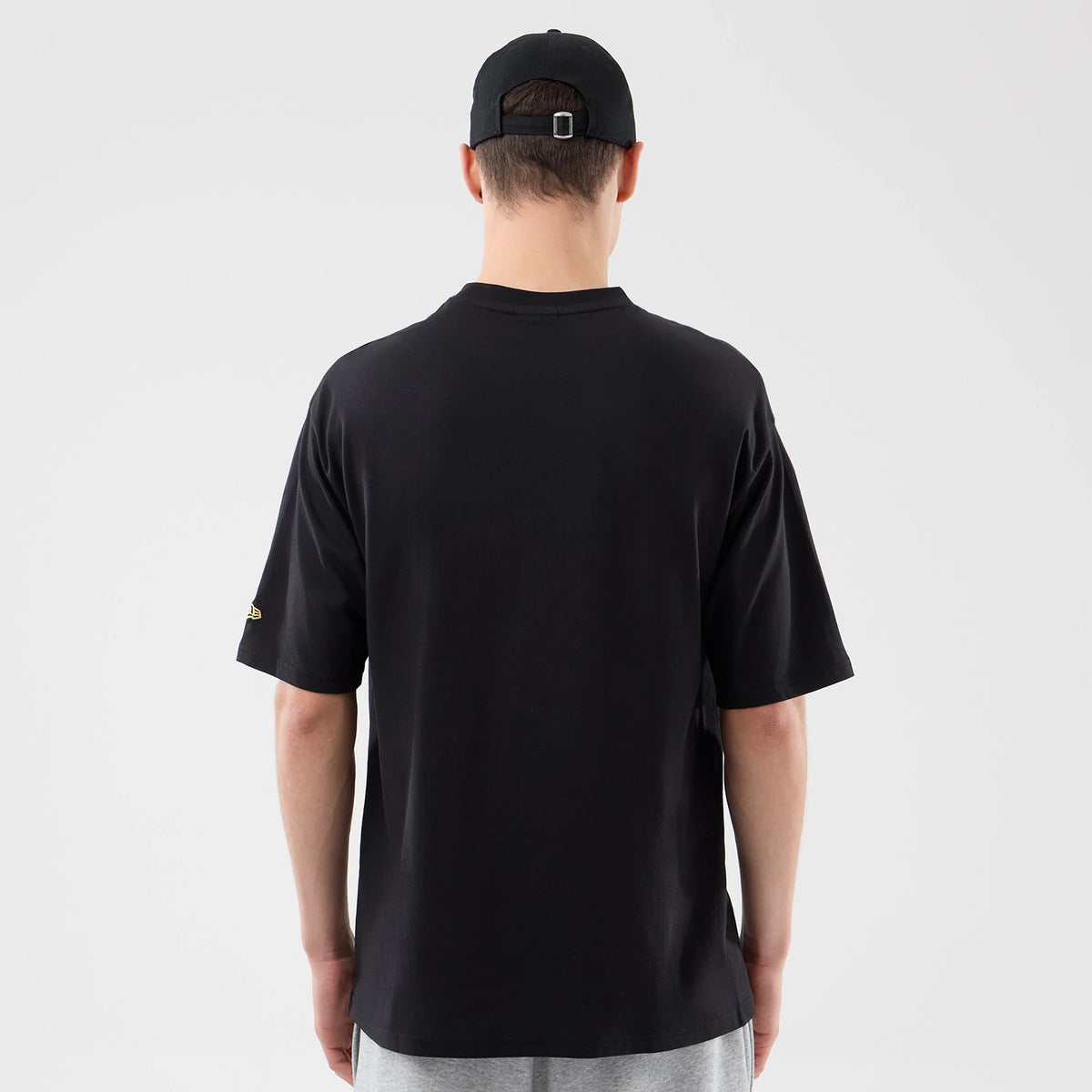 New Era Sticker Lisbon Black Oversized T-Shirt, back view on a model wearing a black cap, showcasing the plain black rear of the oversized tee and a small embroidered New Era logo in gold on the left sleeve.