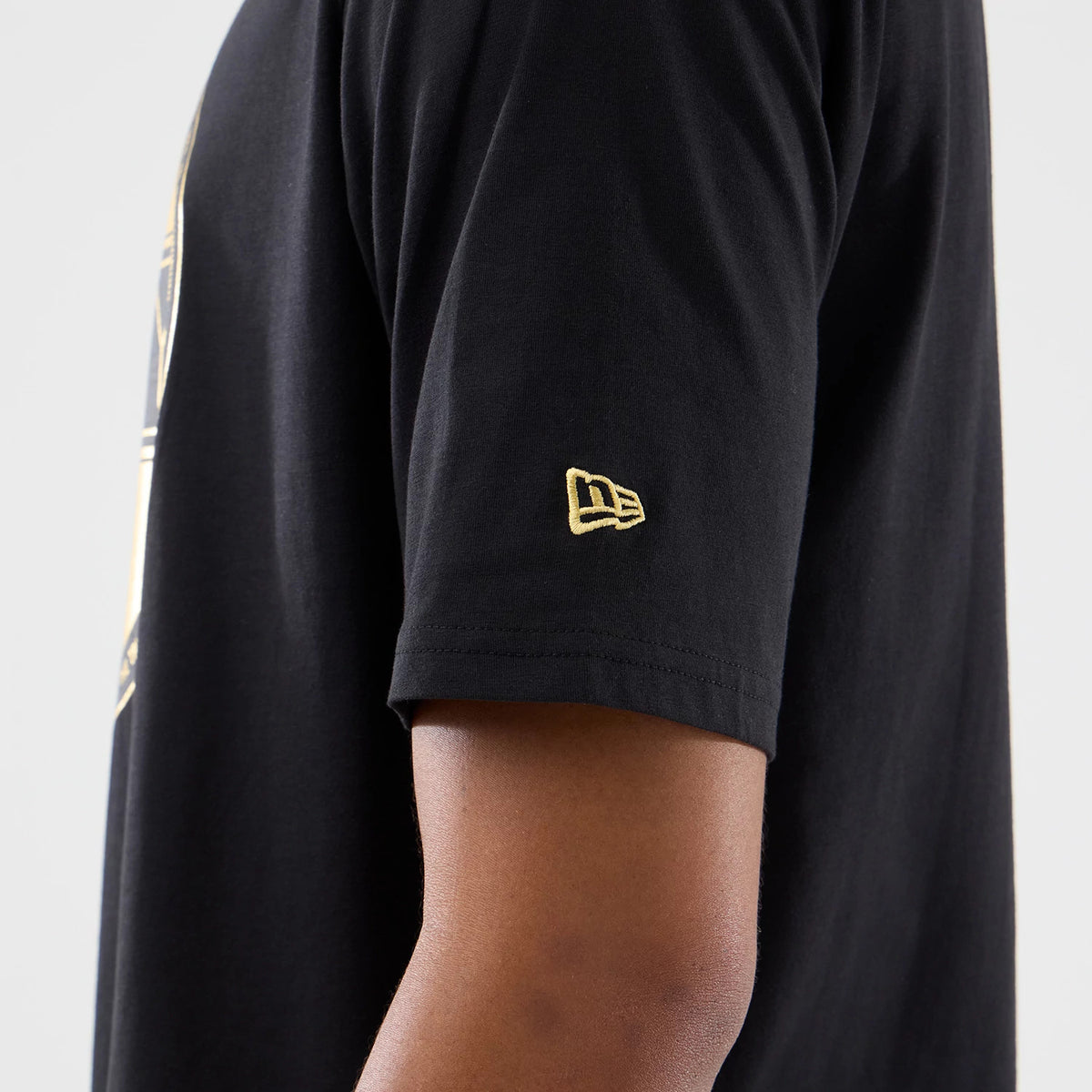 New Era Sticker Madrid Black Oversized T-Shirt, side view on-model featuring a short sleeve with an embroidered gold New Era logo near the hem, part of a larger graphic along the side of the garment.