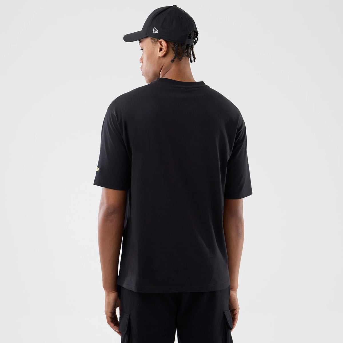 New Era Sticker Madrid Black Oversized T-Shirt, back view on a model wearing a black cap, showing the plain black rear with a small yellow embroidered logo on the left sleeve and loose short sleeves extending to the elbows.