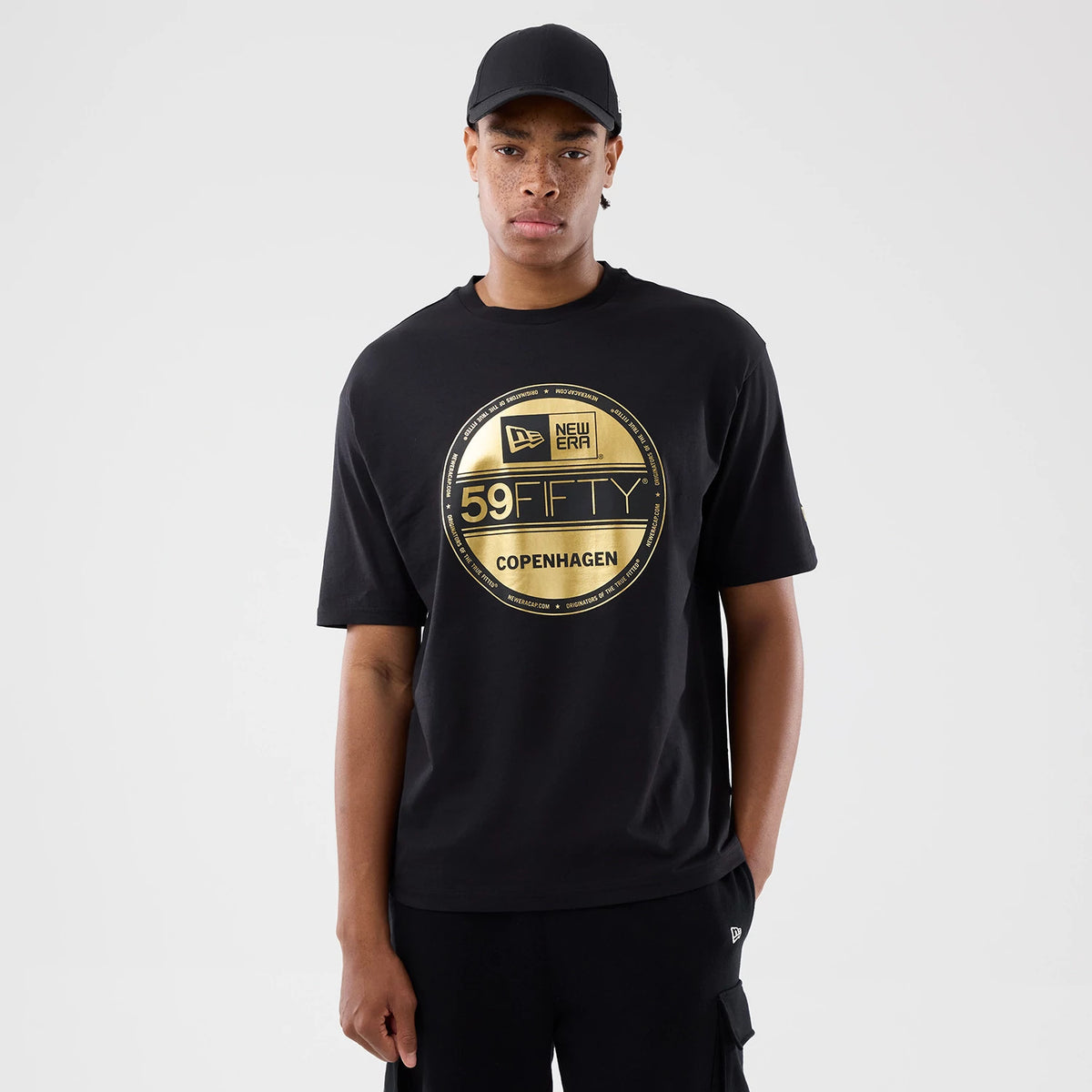 New Era Sticker Copenhagen Black Oversized T-Shirt, front view on a model showing a large gold printed circular design featuring the New Era logo, 59FIFTY and Copenhagen text, with relaxed short sleeves and a straight hem.