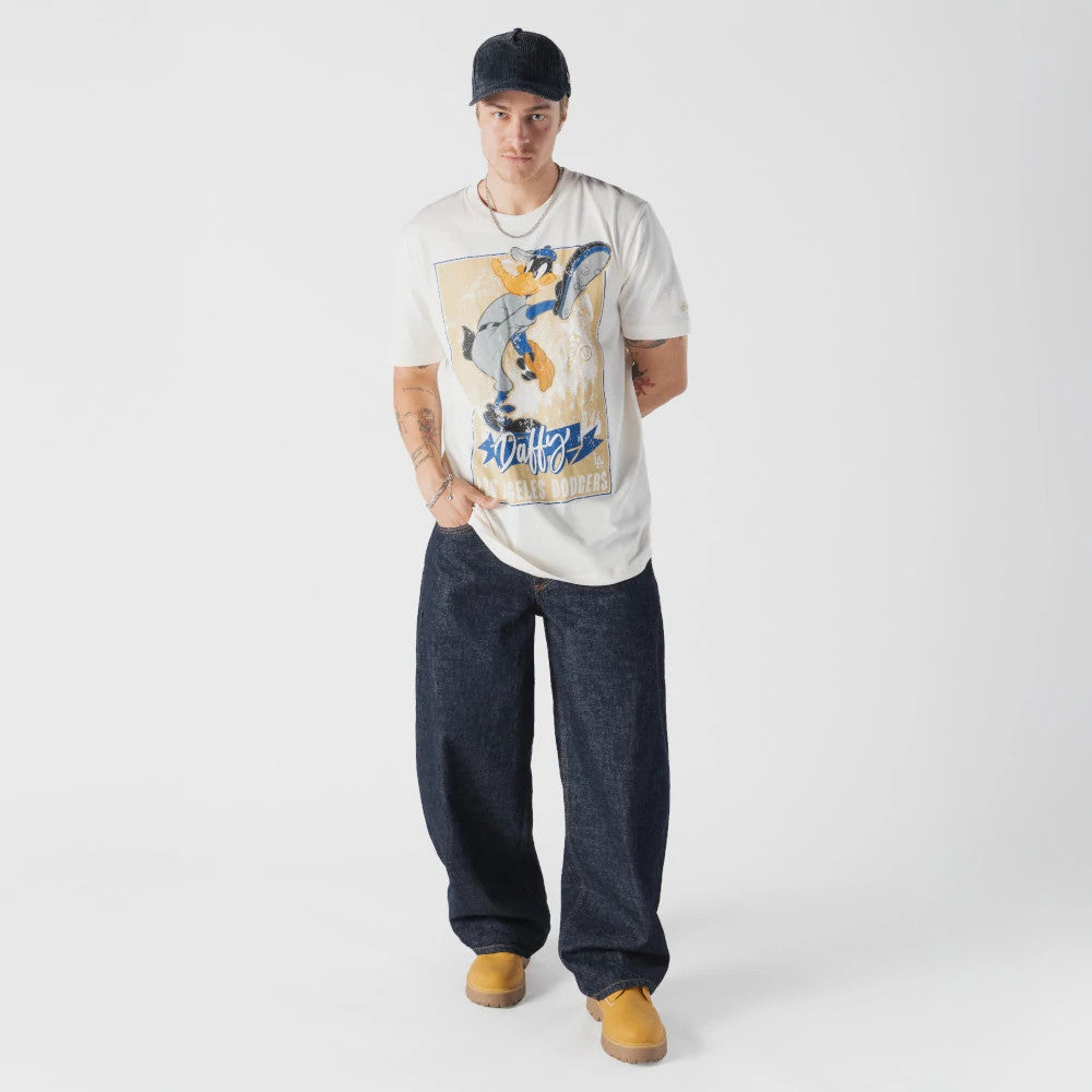 New Era LA Dodgers MLB X Looney Tunes Cream T-Shirt, front view featuring a large printed graphic of Daffy Duck in action on the centre, LA Dodgers text below, worn by a model styled with loose dark jeans and tan boots for a casual look.