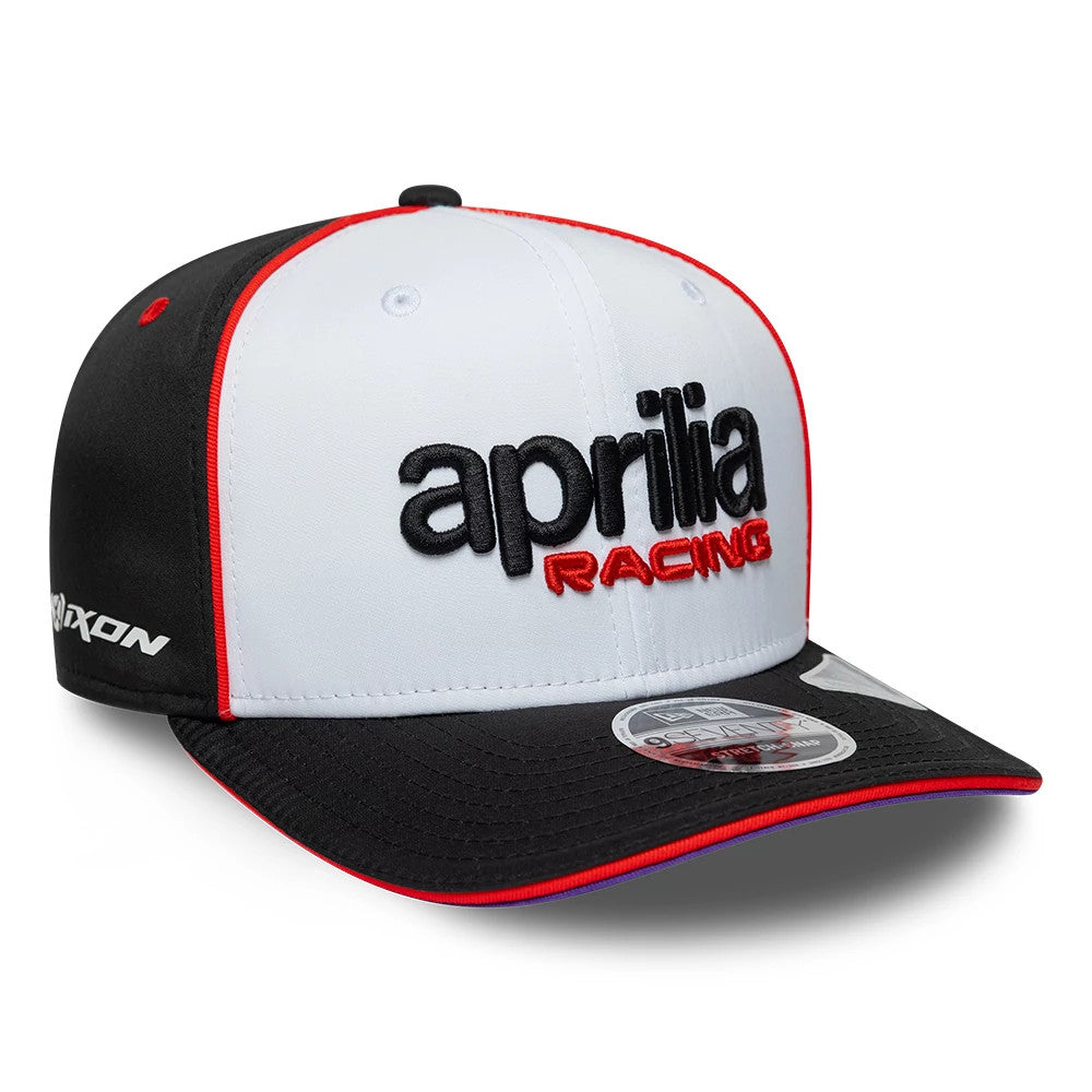 Aprilia Team White New Era 9SEVENTY Stretch Snap Adjustable Cap, three-quarter view showing aprilia RACING in black and red embroidery on the white front panel, contrasting red trim, and a black visor with a 9SEVENTY size sticker.