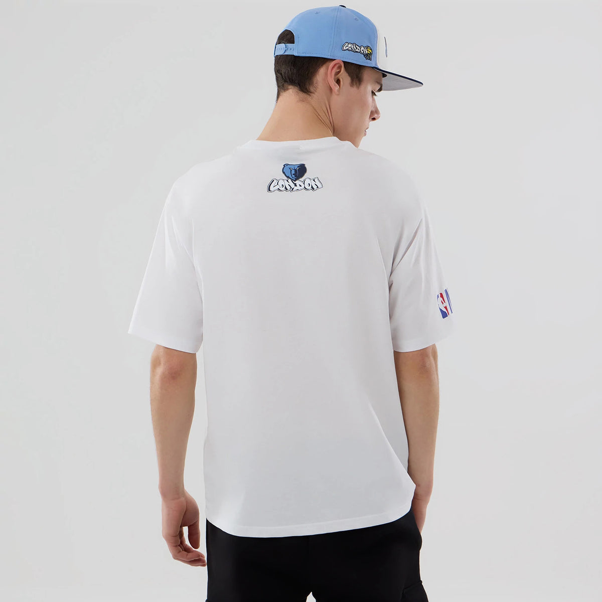 New Era Memphis Grizzlies NBA London Game 2026 White Oversized T-Shirt, back view showing a small blue and black graphic near the collar, NBA logo embroidered on the left sleeve, worn by a model with a blue and black cap.