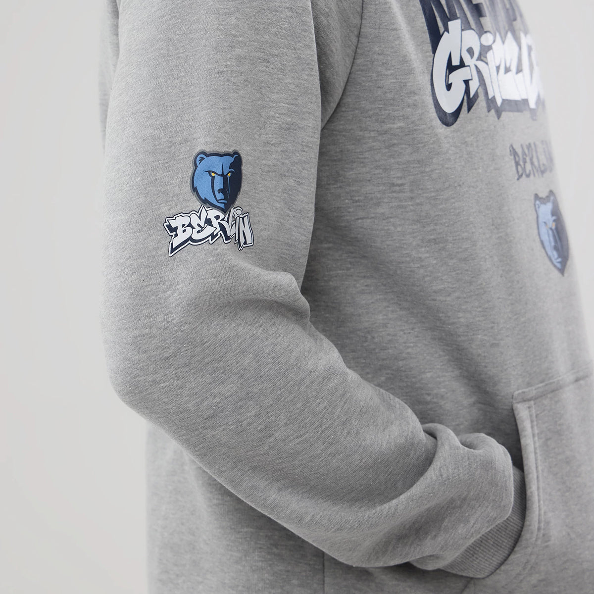 New Era Memphis Grizzlies NBA Berlin Game 2026 Grey Pullover Hoodie, side view showing the grey hoodie with the Grizzlies logo printed on the sleeve, Berlin lettering beneath, and view of the chest graphic with kangaroo pocket.