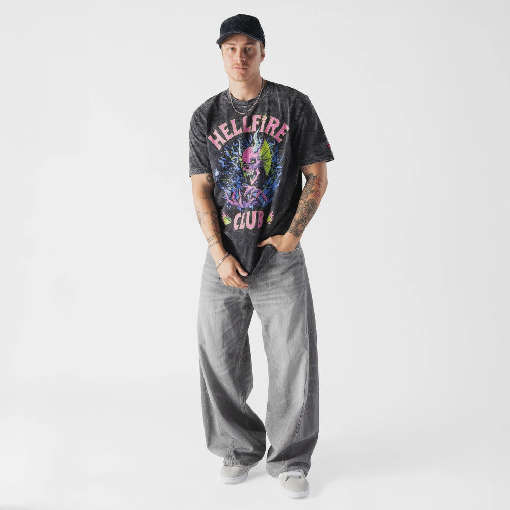 New Era Netflix Stranger Things Hell Fire Club Black T-Shirt, front view on-model showing a graphic with a demonic face in the centre, surrounded by flames and stylised lettering reading Hellfire Club, paired with grey trousers and white trainers.