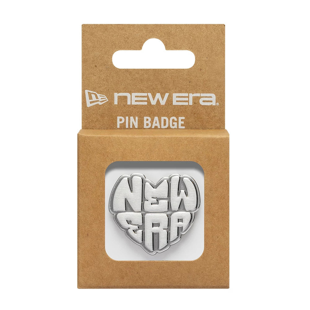 New Era Heart Silver Pin Badge, front view inside packaging showing a heart-shaped silver badge with raised New Era lettering, packaged in a brown cardboard box with a cut-out window and printed New Era branding on the top flap.