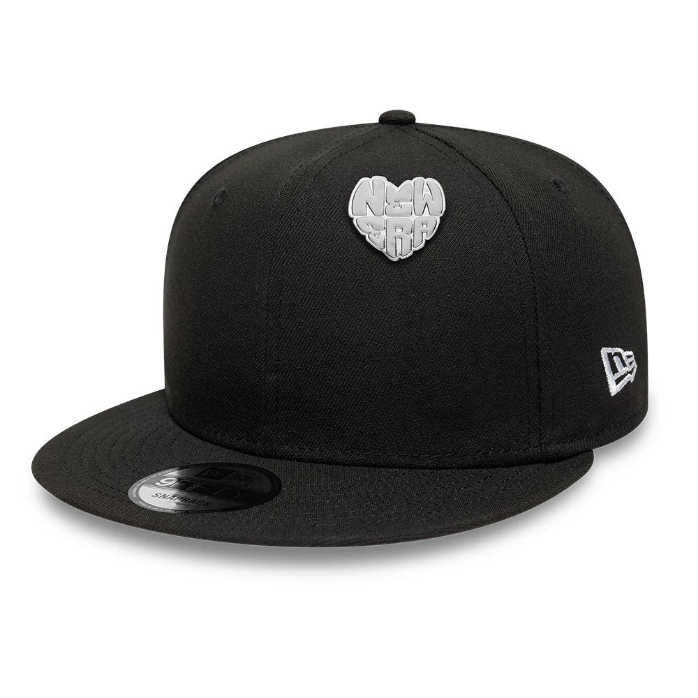 New Era Heart Silver Pin Badge, front view on a black cap showcasing a raised silver heart-shaped badge with lettering on the crown, accompanied by an embroidered white New Era logo on the side and a flat black visor.