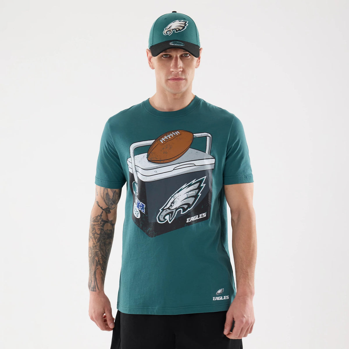 New Era Philadelphia Eagles NFL Training 2026 Dark Green T-Shirt, front view on a model wearing a matching cap, showing a large graphic of a cooler with a football and the Eagles logo, accompanied by embroidered New Era and Eagles logos near the hem.