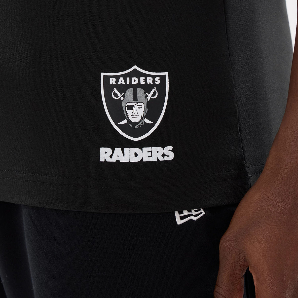 New Era Las Vegas Raiders NFL Training 2026 Black T-Shirt, front view showing a white printed Raiders shield logo and text at the hem, worn by a model also wearing dark trousers with the New Era logo just above the waistband.