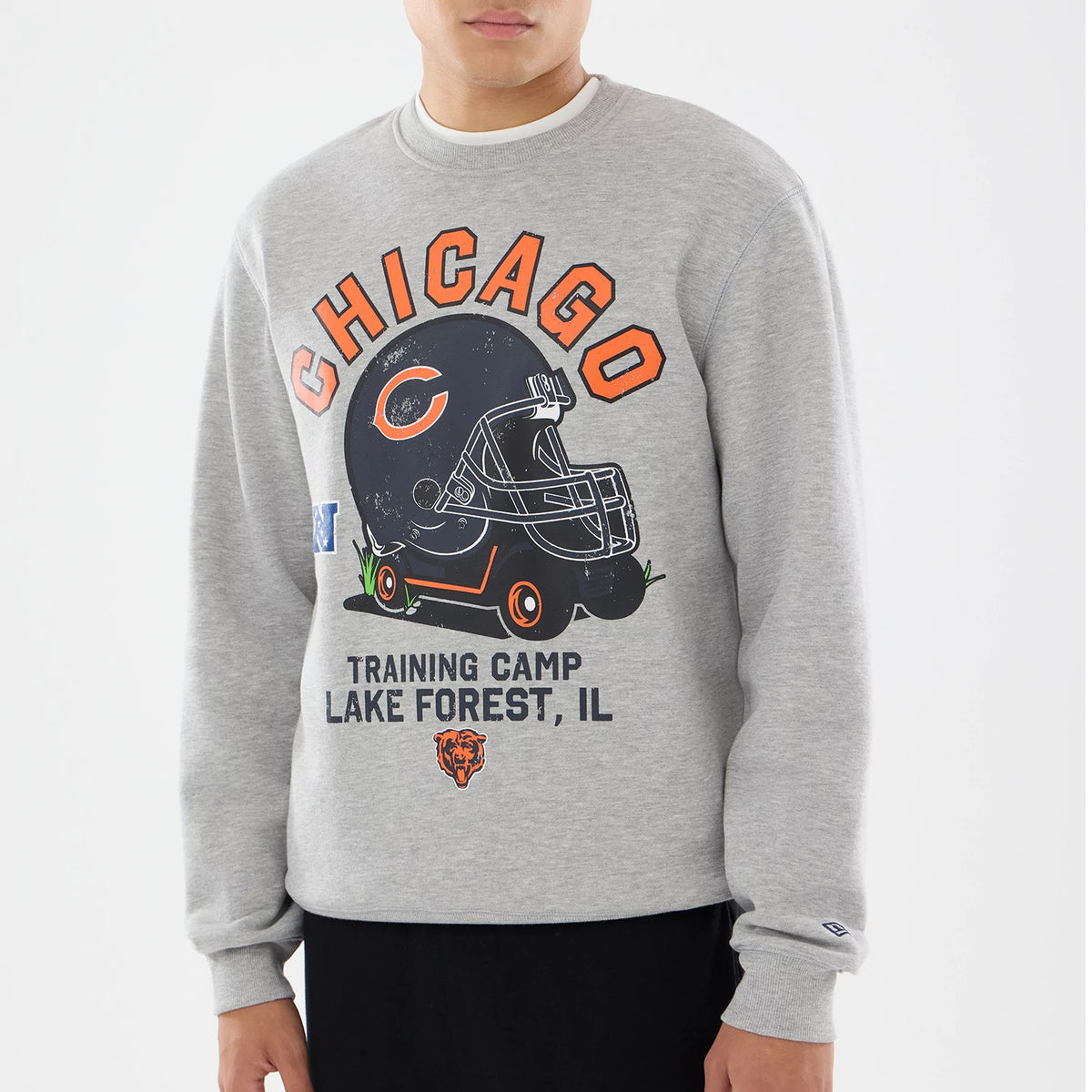 New Era Chicago Bears NFL Training 2026 Grey Crew Neck Sweater, front view on a model showing orange Chicago lettering above a large vintage helmet graphic with the Bears logo, and detailed Training Camp Lake Forest, IL text below.