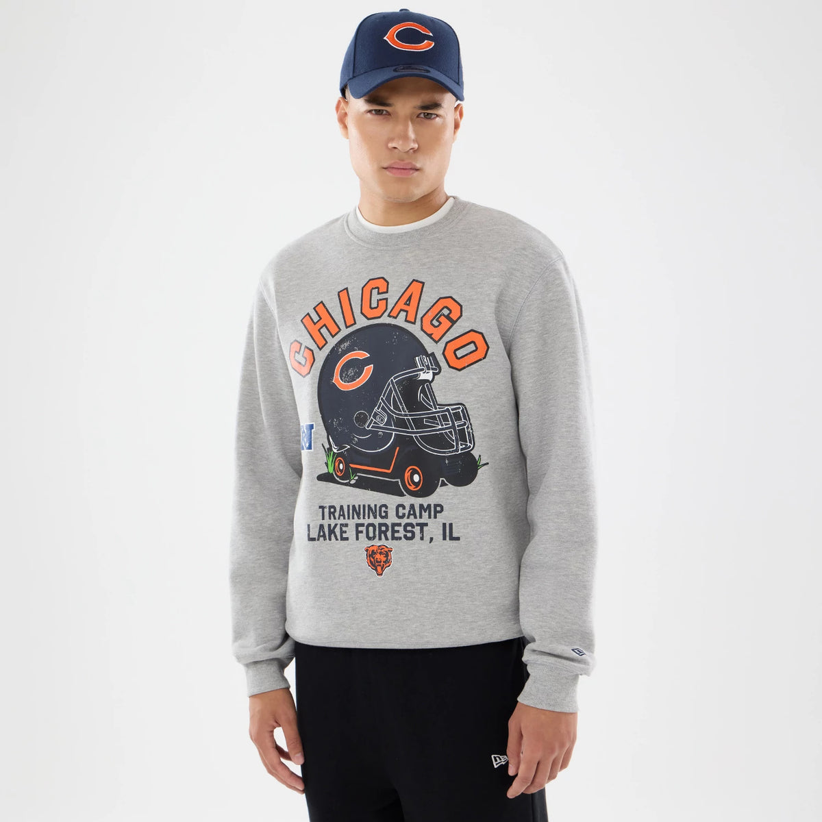 New Era Chicago Bears NFL Training 2026 Grey Crew Neck Sweater, front view on a model with a navy cap, featuring orange Chicago lettering above a detailed graphic of a football helmet, and a New Era flag logo on the sleeve.