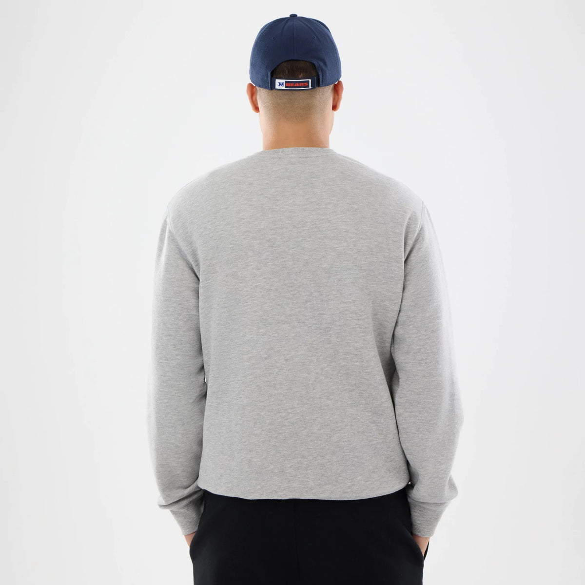 New Era Chicago Bears NFL Training 2026 Grey Crew Neck Sweater, rear view on a model wearing a navy cap, showing the sweater's plain grey back with long sleeves, ribbed cuffs, and a relaxed fit, paired with black trousers.