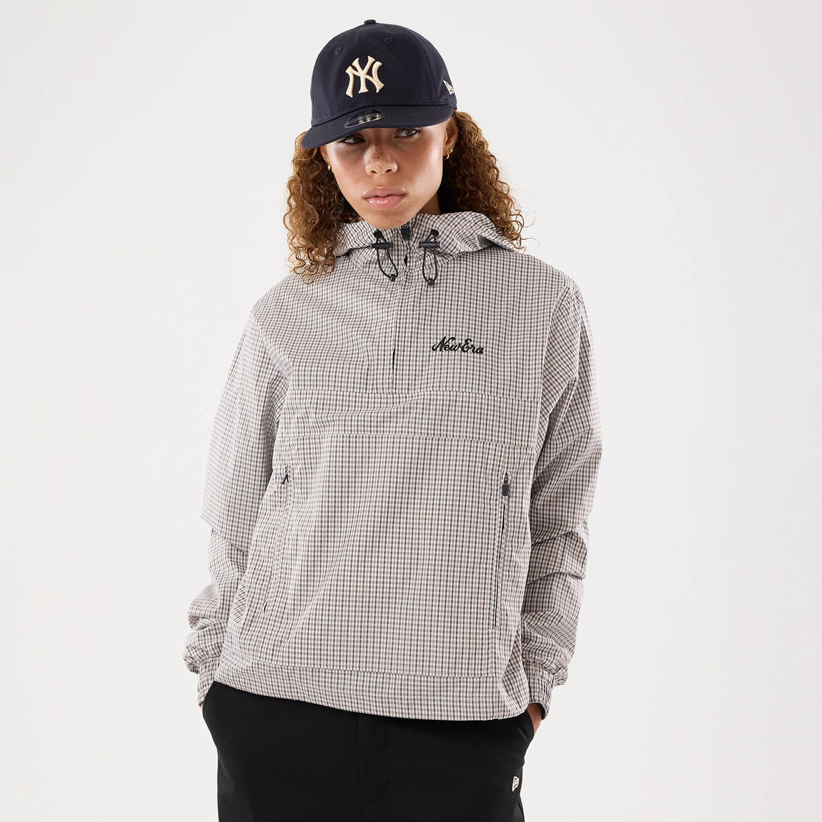 Womens New Era Check Beige Oversized Windbreaker Jacket, front view on a model wearing a black cap, featuring a beige and white check pattern, embroidered New Era logo on the chest, and zippered pockets on the front.