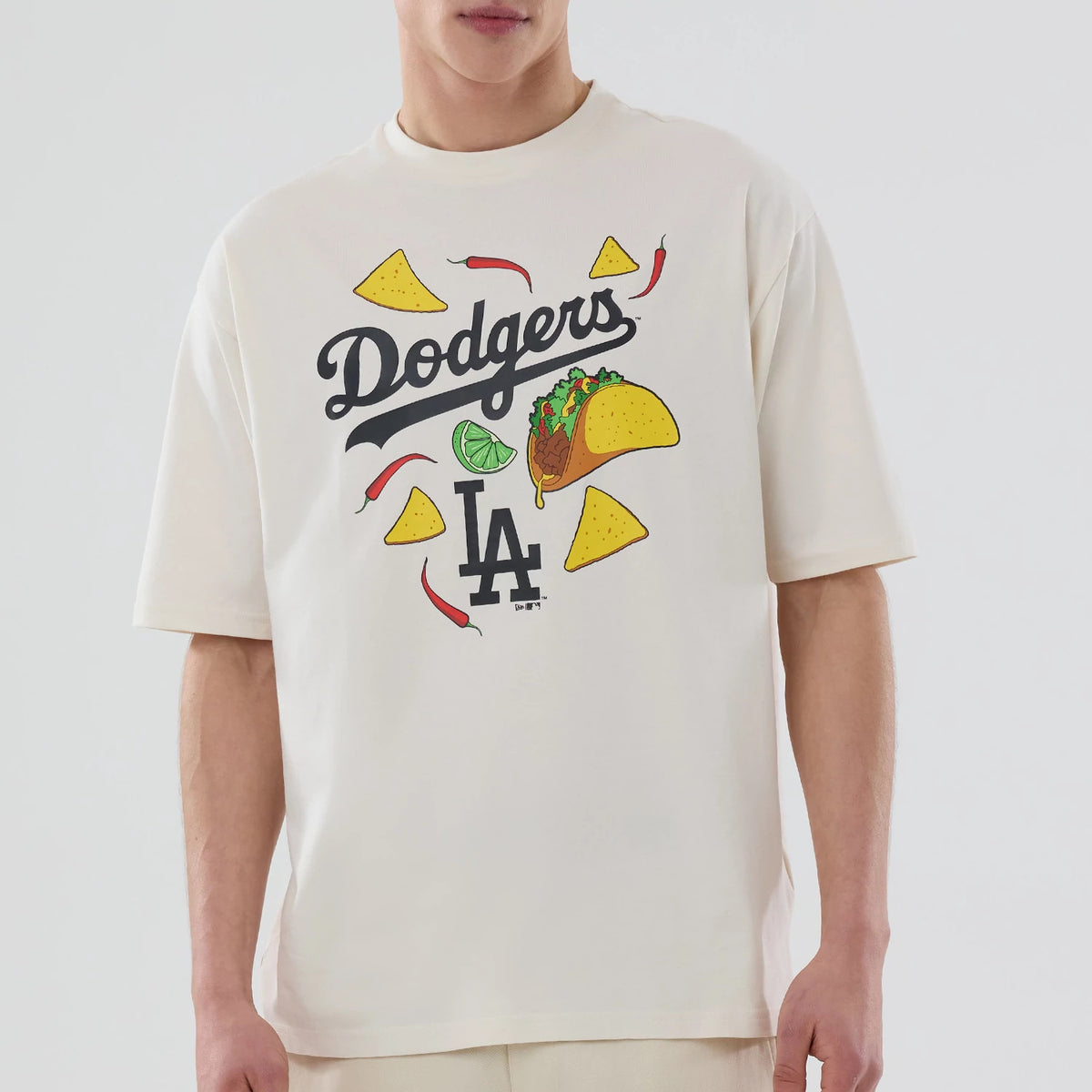 New Era LA Dodgers MLB Food Graphic Open White Oversized T-Shirt, front view displaying colourful graphics including tacos, chillies, and tortilla chips surrounding the Dodgers script and the LA logo, on a model with neutral trousers.