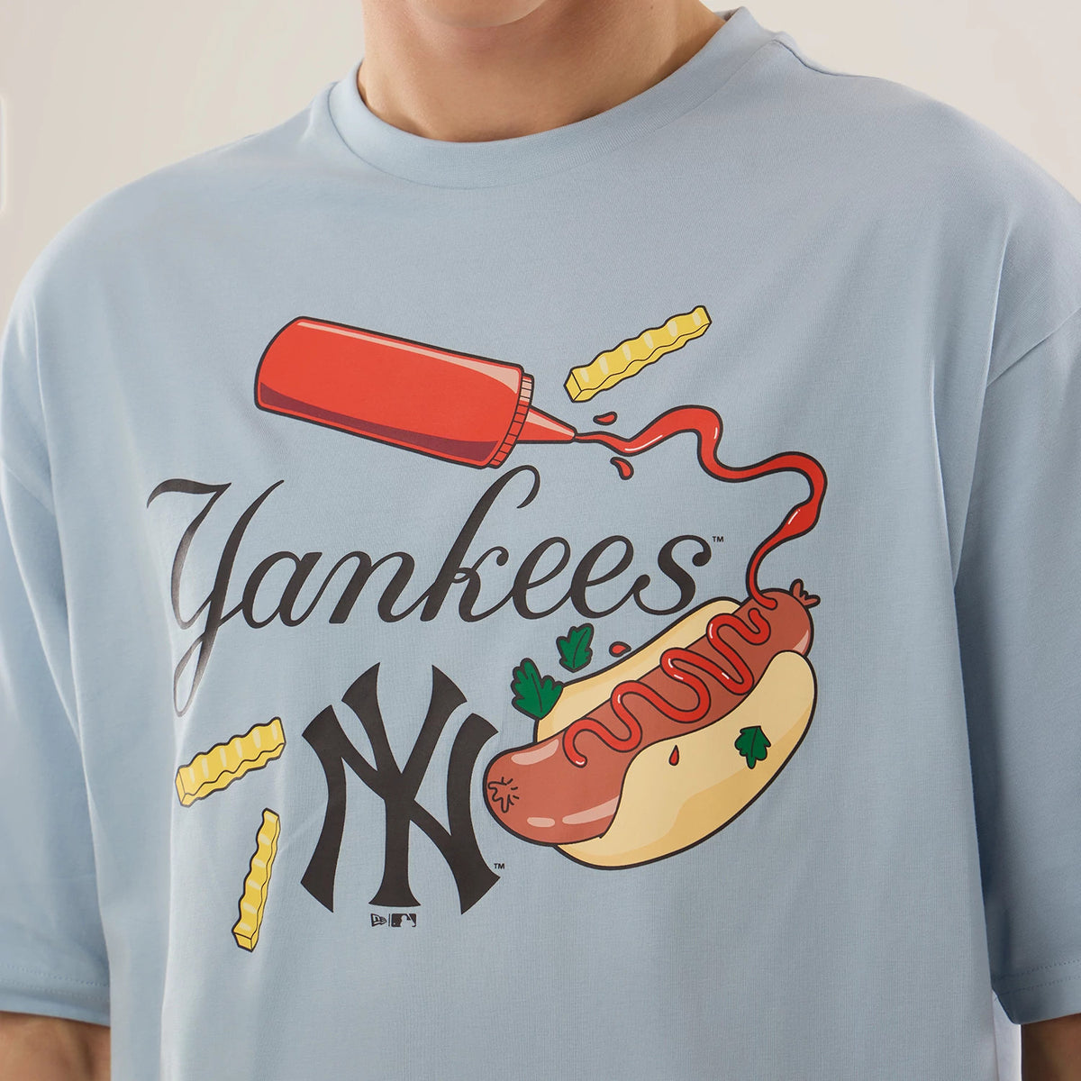 New Era New York Yankees MLB Food Graphic Pastel Blue Oversized T-Shirt, front view on-model showing a graphic of a hot dog with ketchup and crinkle-cut fries, the word Yankees in script across the chest above a black printed NY Yankees logo.