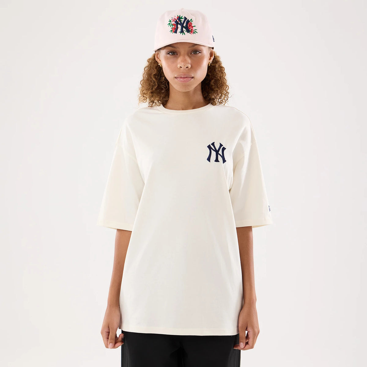 New Era New York Yankees Womens MLB Graphic Open Cream T-Shirt, front view on a model featuring a navy embroidered NY logo on the left chest, with short sleeves and relaxed fit, paired with a matching cap with a central embroidered NY logo.