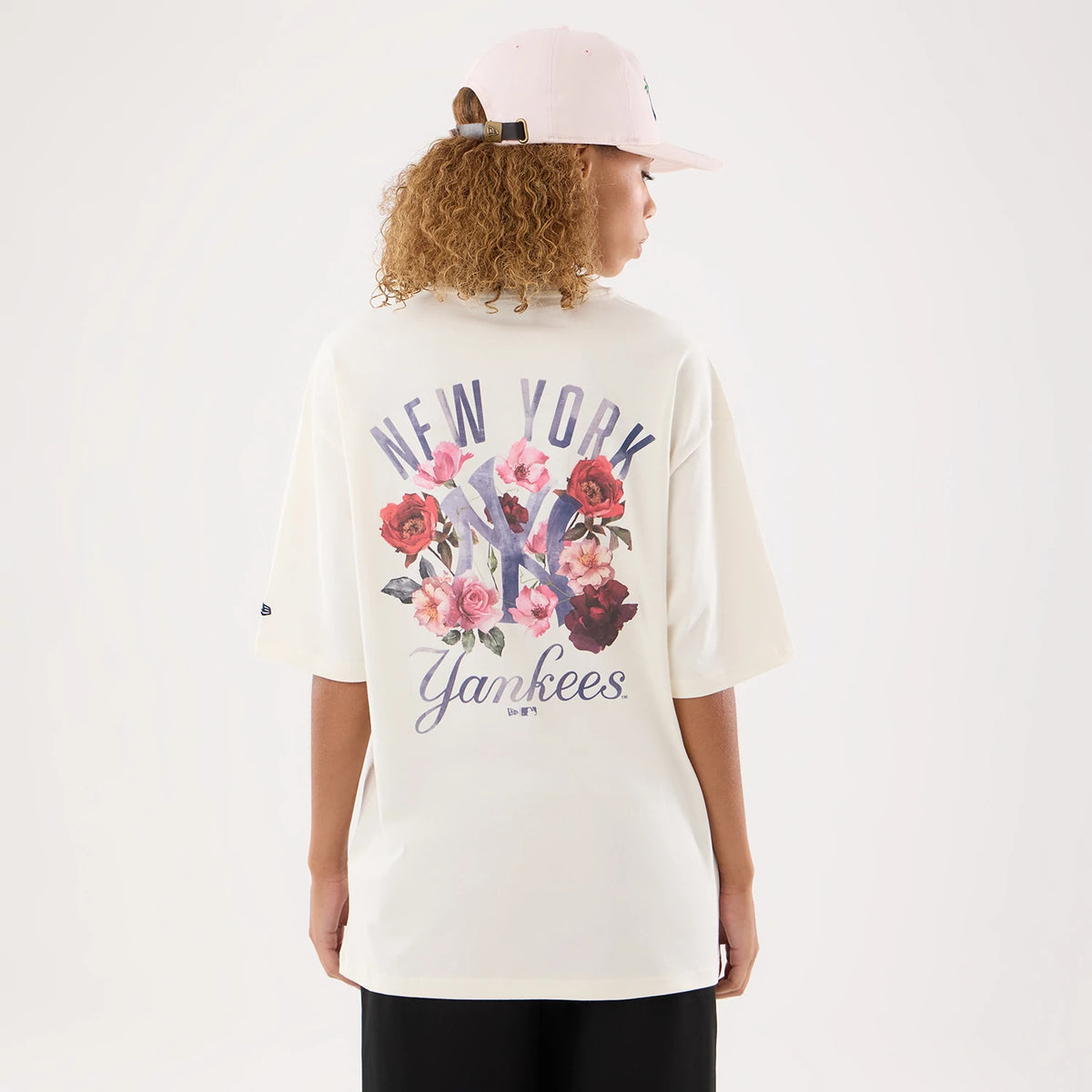 New Era New York Yankees Womens MLB Graphic Open Cream T-Shirt, back view on a model with a pastel pink cap, displaying large floral graphics surrounding a central NY logo, with New York lettering above and Yankees written below in blue script.