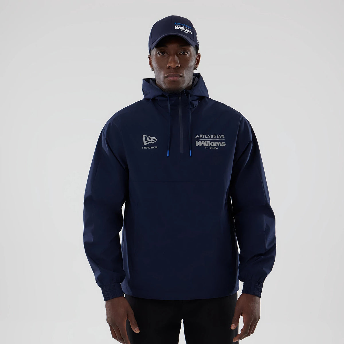 New Era Williams Navy Windbreaker Jacket, front view on-model displaying an embroidered New Era logo on the right chest, Atlassian and Williams F1 Team embroidery on the left chest, and a drawstring hood, styled with a matching navy cap.