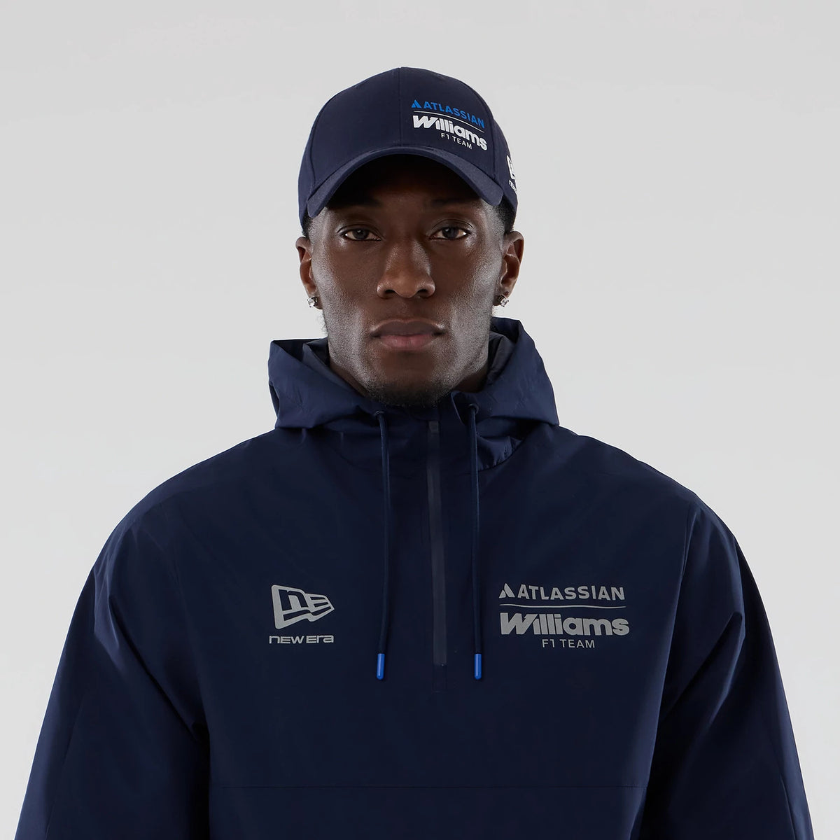 New Era Williams Navy Windbreaker Jacket, front view on-model showing the navy jacket with an embroidered New Era logo on the left chest, Atlassian Williams F1 Team branding on the right chest and a matching cap styled with the outfit.