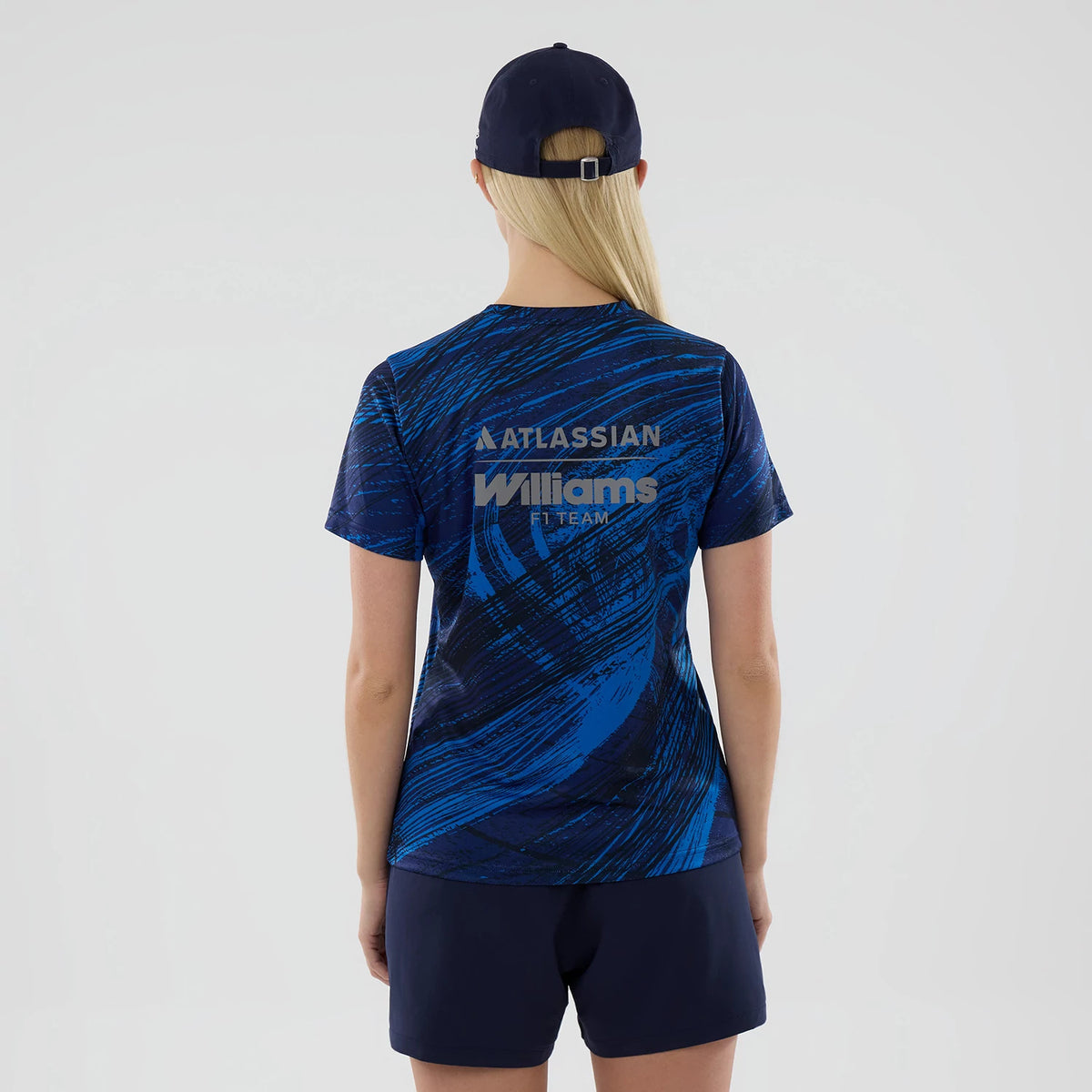 New Era Williams Womens Navy Active T-Shirt, back view on a model wearing matching navy cap, featuring blue swirl graphics and raised Williams F1 Team lettering in the centre, with Atlassian branding across the shoulders.