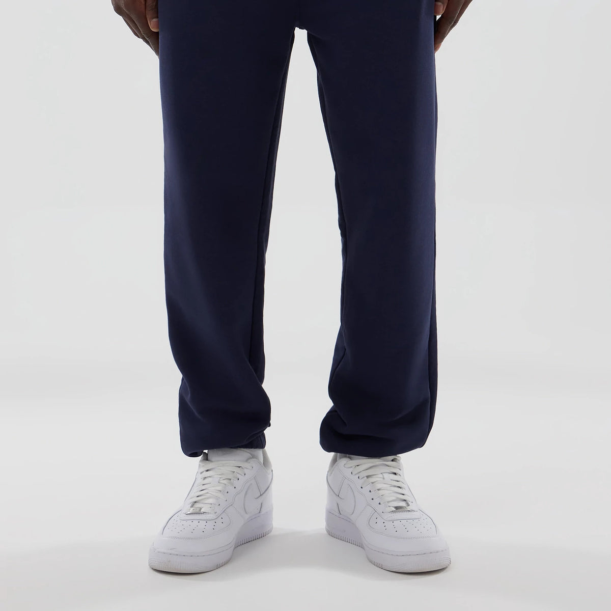 New Era Williams Navy Joggers, front view showing the clean navy fabric with a straight leg fit, worn with plain white trainers and reaching just above the ankle, while a model's fingers rest gently alongside the seams.