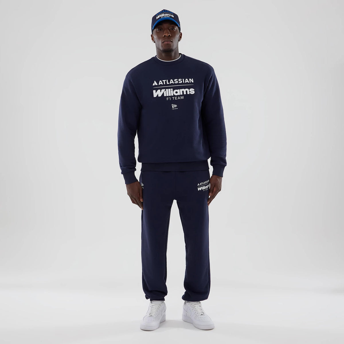 New Era Williams Navy Joggers, on-model view with a matching navy sweatshirt featuring white branding on the top left leg, elasticated cuffs, and paired with white trainers for a complete uniform look.