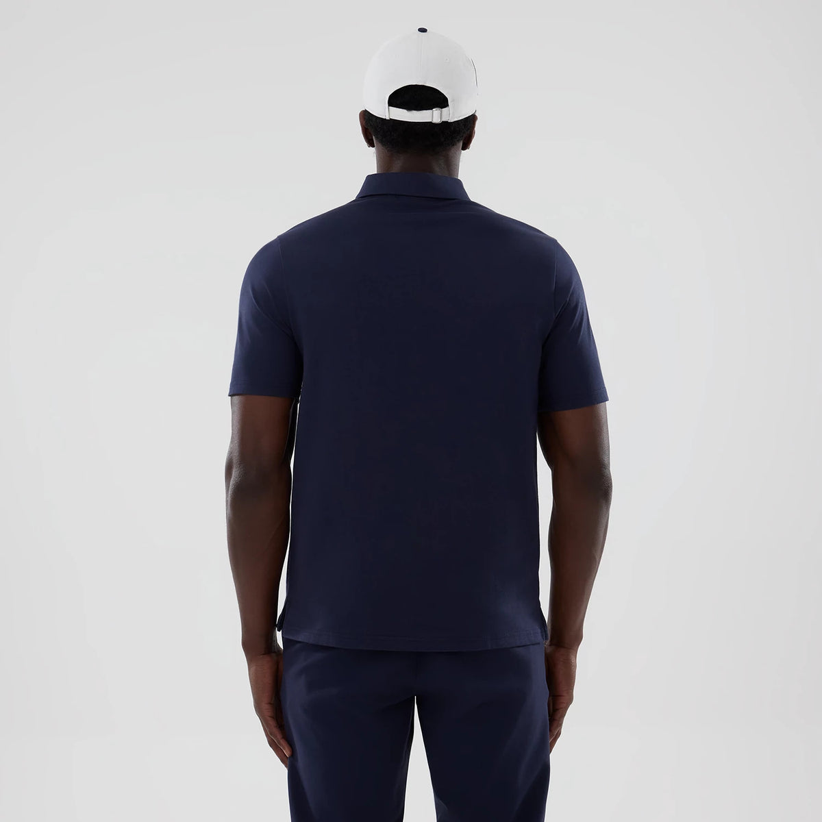New Era Williams Navy Polo T-Shirt, back view on a model showcasing the plain navy fabric with a neat collar and short sleeves, styled with matching navy trousers and a white cap, highlighting the shirt's classic.