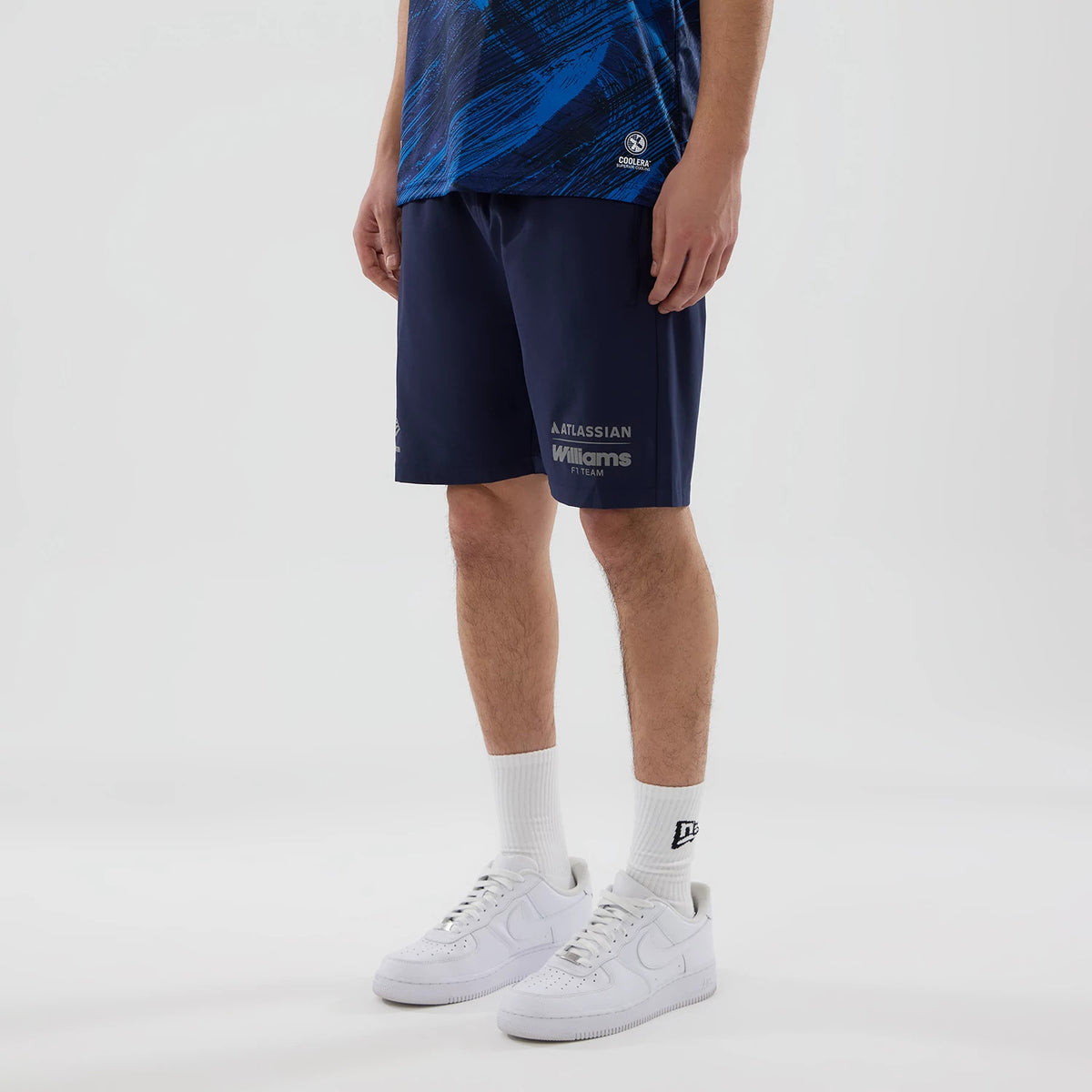 New Era Williams Navy Training Shorts, on-model view showcasing the knee-length navy shorts with light grey printed Atlassian and Williams Racing logos on the front, styled with white trainers and socks featuring an embroidered New Era logo.