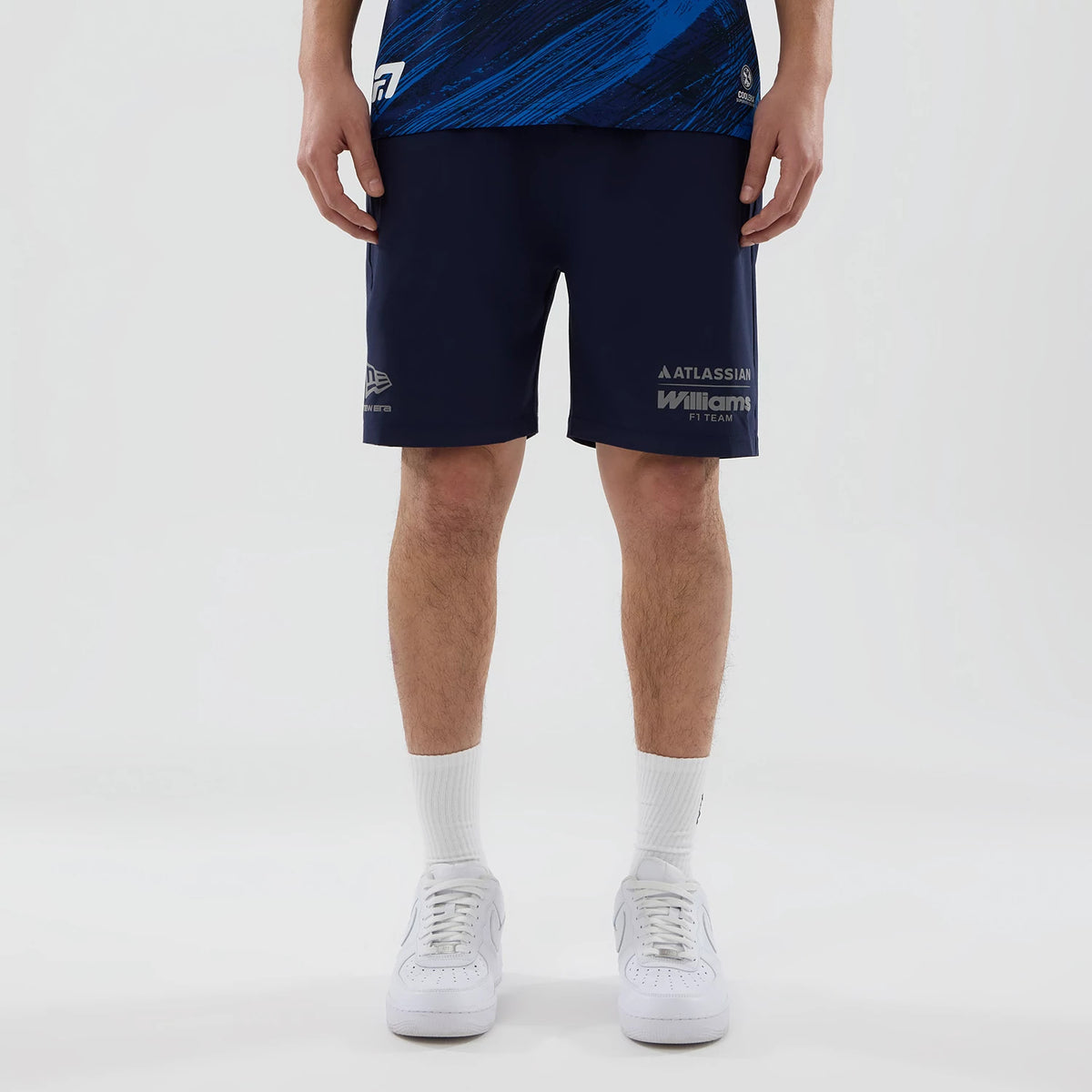 New Era Williams Navy Training Shorts, front view on-model showing printed Williams F1 Team logo on the lower right leg, embroidered New Era logo on the lower left leg, and a navy body with a relaxed fit styled with white trainers and socks.