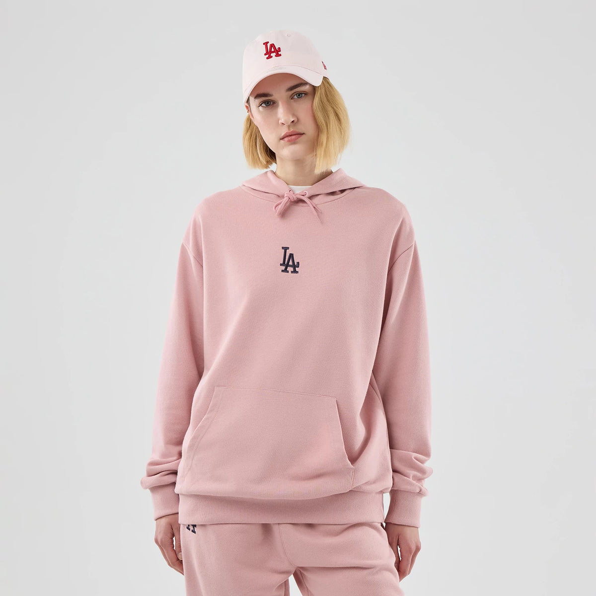 New Era Dodgers MLB Midi League Essential Pastel Pink Oversized Pullover Hoodie, front view on-model the pink hoodie with an embroidered LA logo on the chest, large front pocket, styled with matching pastel pink trousers and a cap.