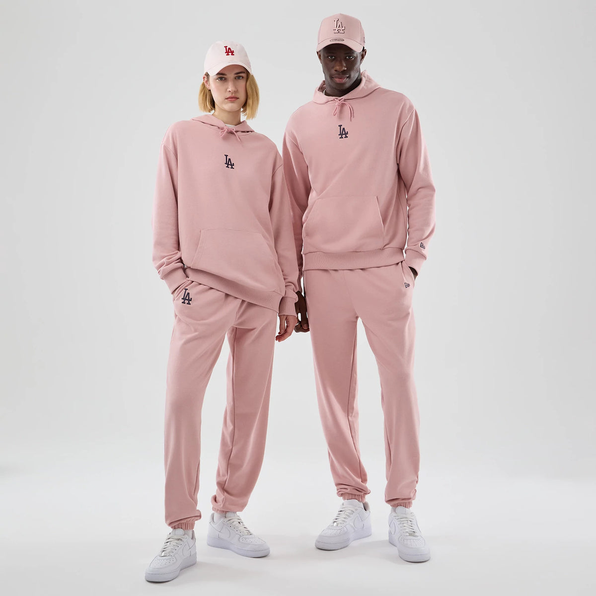 New Era Dodgers MLB Midi League Essential Pastel Pink Oversized Pullover Hoodie, front view on a model showcasing the relaxed fit with an embroidered LA logo on the chest, paired with matching pink trousers and a cap with a raised LA logo.