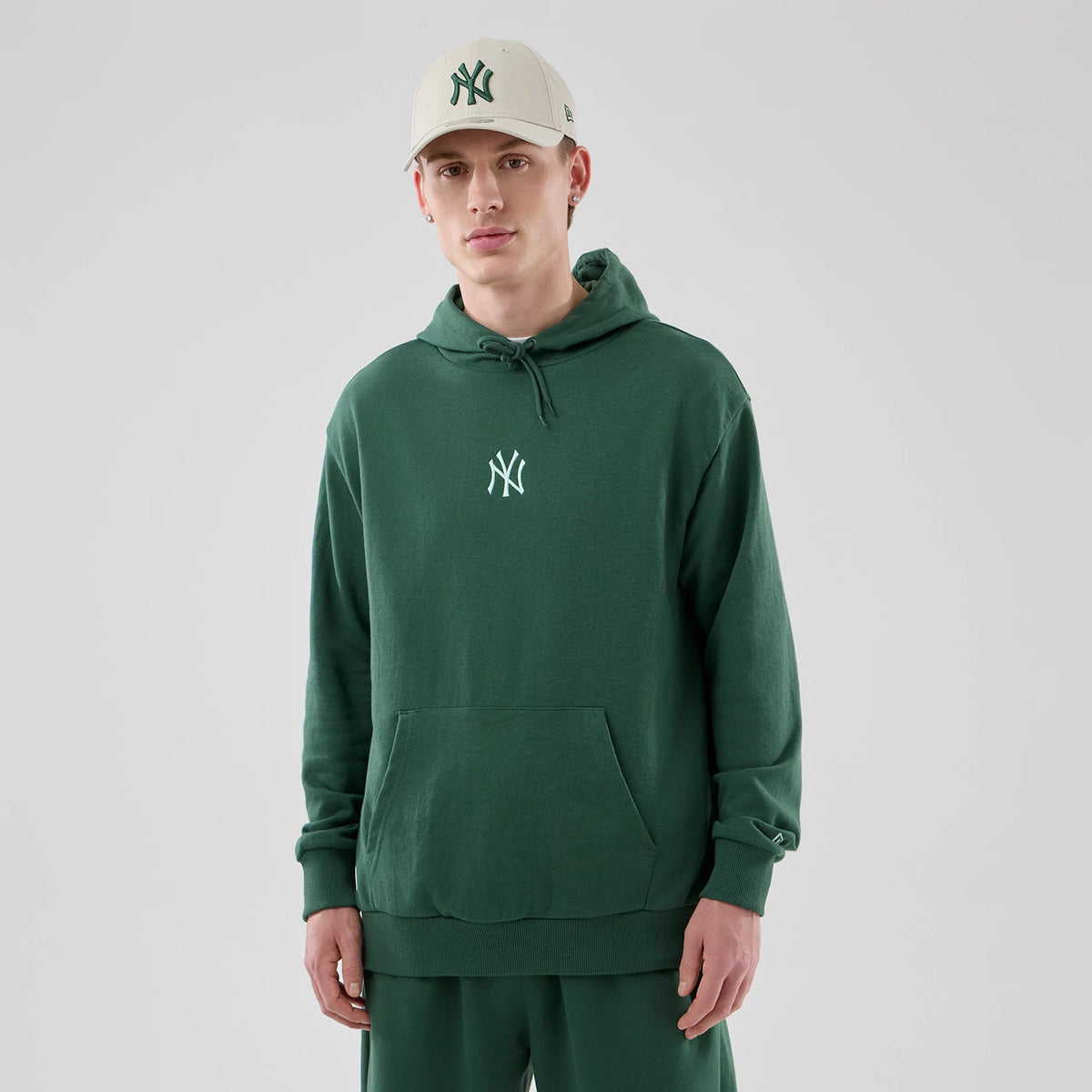 New Era Yankees MLB Midi League Essential Dark Green Oversized Pullover Hoodie, front view on a model also wearing a matching beige cap, featuring a white embroidered Yankees logo at the centre chest and a spacious front kangaroo pocket.