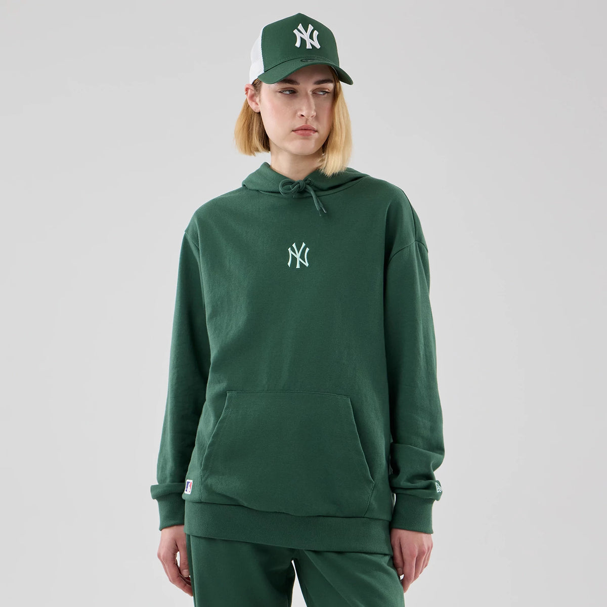 New Era Yankees MLB Midi League Essential Dark Green Oversized Pullover Hoodie, front view on a model with loose green trousers, featuring a white embroidered NY logo on the chest, a drawstring hood, and a matching logo cap worn by the model.