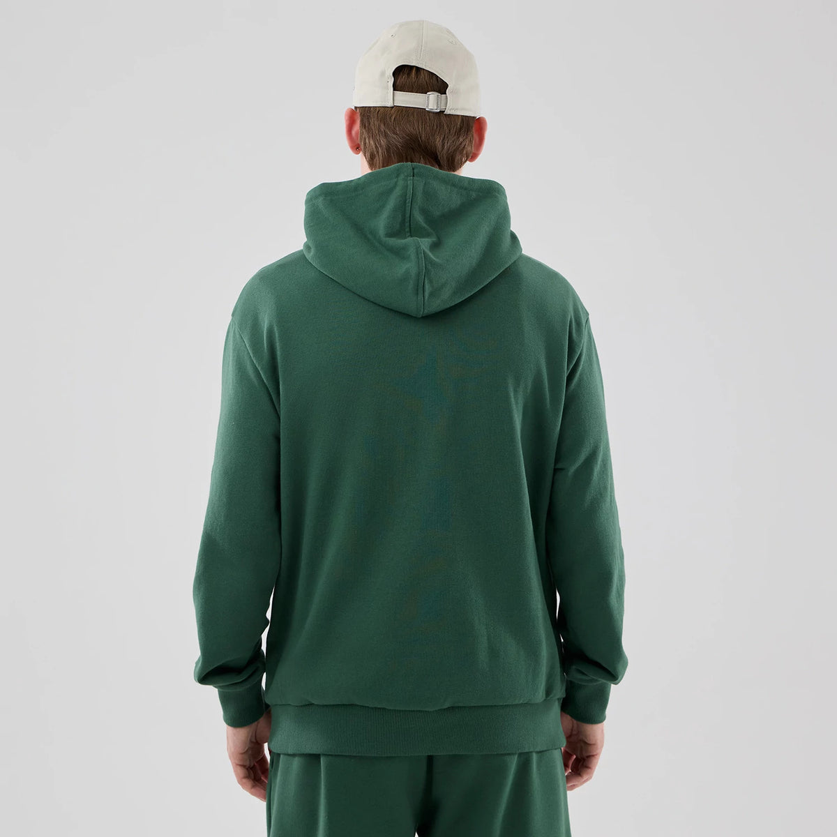 New Era Yankees MLB Midi League Essential Dark Green Oversized Pullover Hoodie, back view on a model showcasing the relaxed fit with a large hood, ribbed hem and cuffs, and worn over matching dark green trousers paired with a plain light cap.