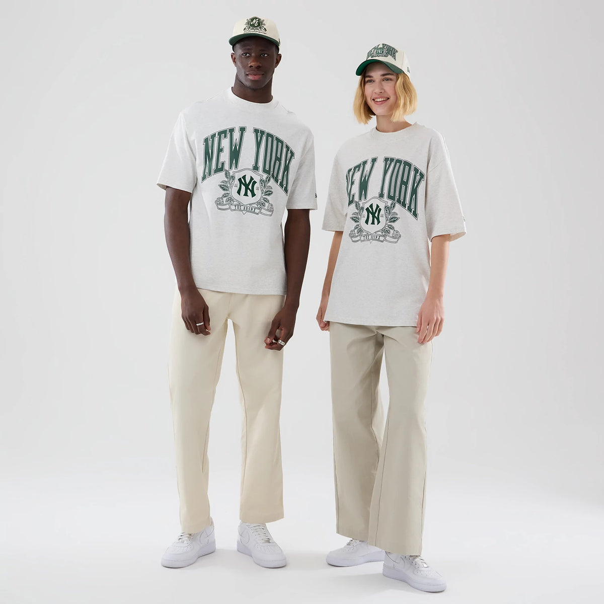 New Era New York Yankees MLB Arch Graphic Pastel Grey Oversized T-Shirt, front view on-model showing large green arching New York lettering across the chest, an embroidered Yankees logo beneath it, styled with beige trousers and white trainers.