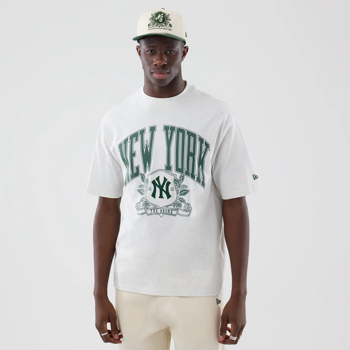 New Era New York Yankees MLB Arch Graphic Pastel Grey Oversized T-Shirt, front view on a model paired with a matching cap, showing large green New York lettering across the chest, an embroidered New Era logo on the sleeve and a central logo graphic.