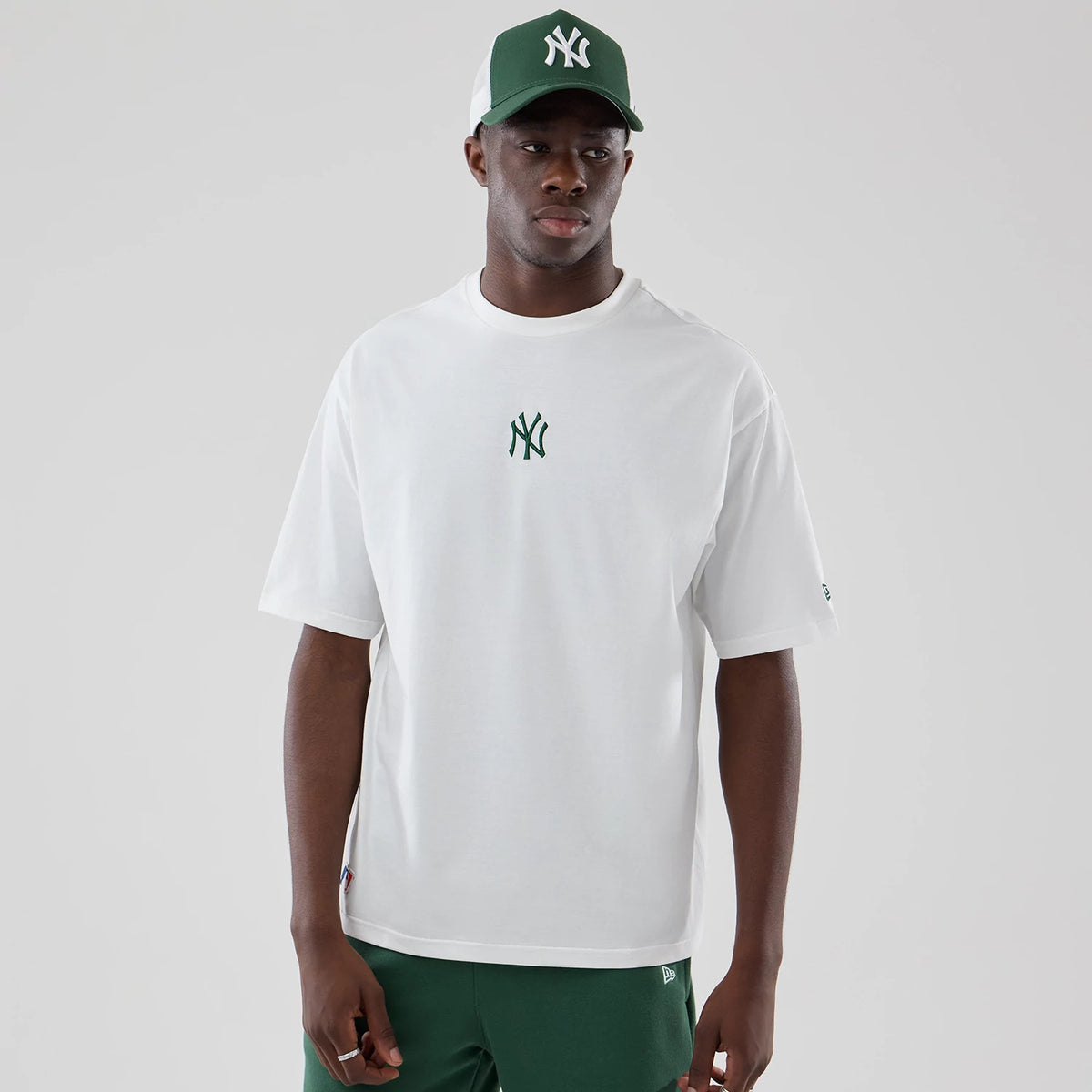 New Era New York Yankees MLB Midi League Essential White Oversized T-Shirt, front view on a model styled with a matching cap, featuring a green embroidered Yankees logo centred on the chest and a small New Era logo on the left sleeve.