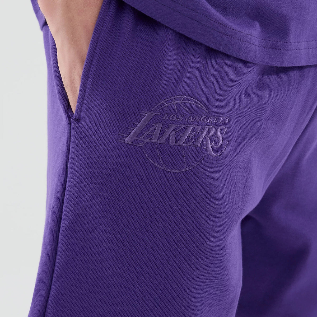 New Era LA Lakers NBA League Essential Purple Oversized Shorts, front view highlighting the purple fabric with an embossed Lakers logo on the left thigh, partially tucked hands in side pockets and the seamless integration into a matching purple top.