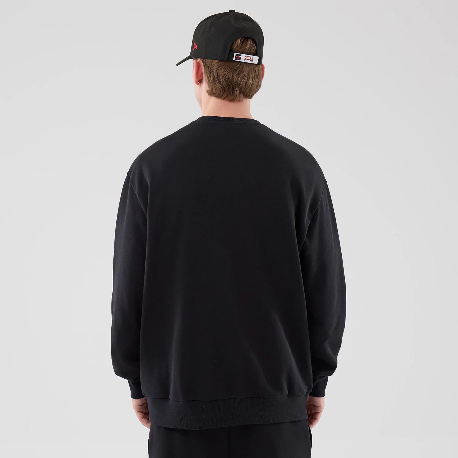 New Era Chicago Bulls NBA League Essential Black Oversized Crew Neck Sweater, back view on a model wearing a black cap, showing the plain black fabric with relaxed shoulders and a loose fit, extending slightly below the waist.