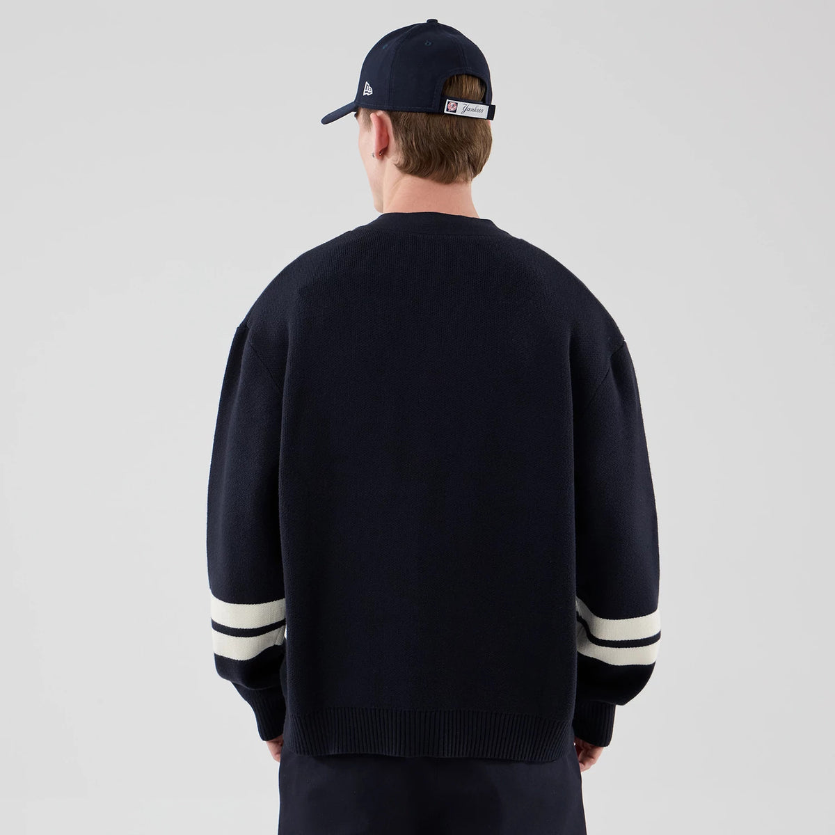 New Era New York Yankees MLB Knit Navy Cardigan, back view on a model also wearing a cap, showing the ribbed hem and cuffs, with two white stripes on each sleeve contrasting against the deep navy knitted fabric.