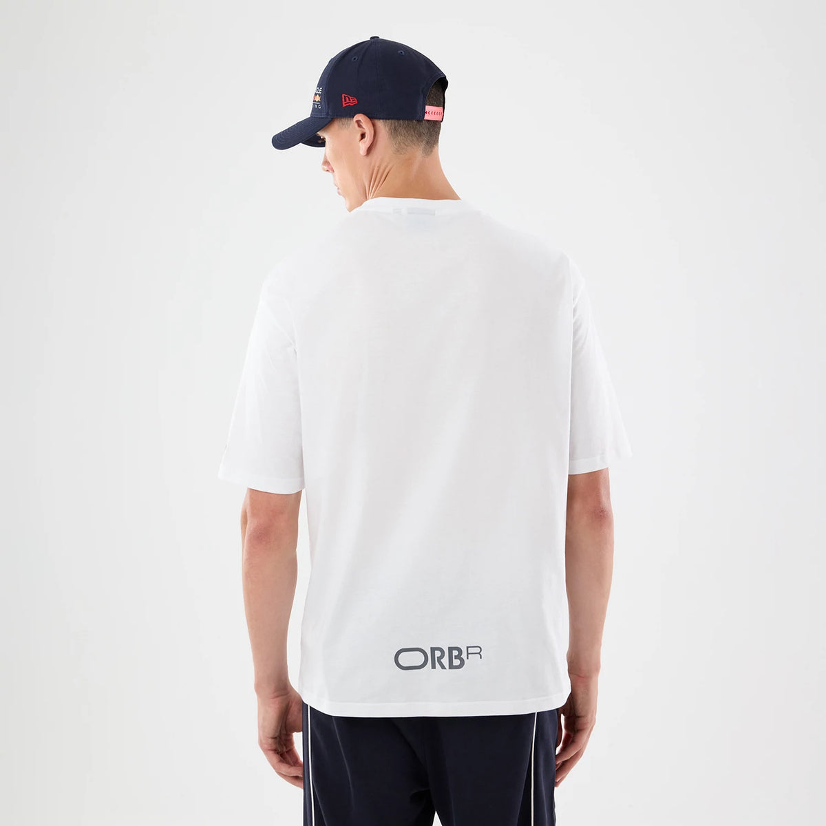 New Era Red Bull Racing Graphic White Oversized T-Shirt, back view on a model wearing a dark cap, showing the ORBR lettering in black near the hem, short sleeves and a clean white back without additional graphics or designs.