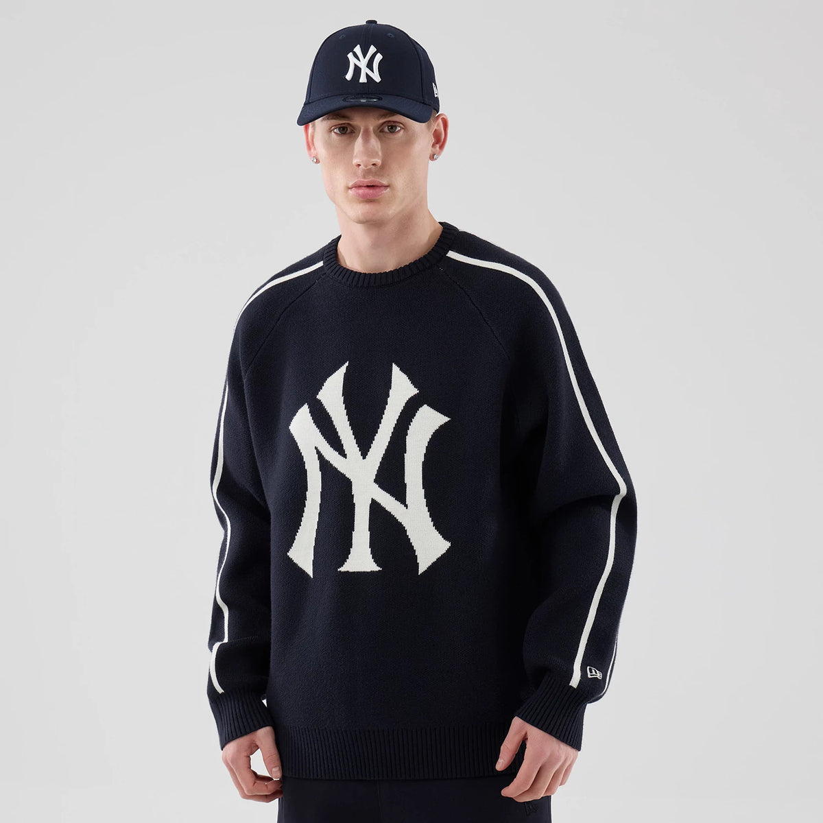 New Era New York Yankees MLB Knit Navy Knitted Sweater, on-model front view with a large white NY logo knitted on the chest, white stripes running down each sleeve, and a crew neck design, styled with a matching navy cap.