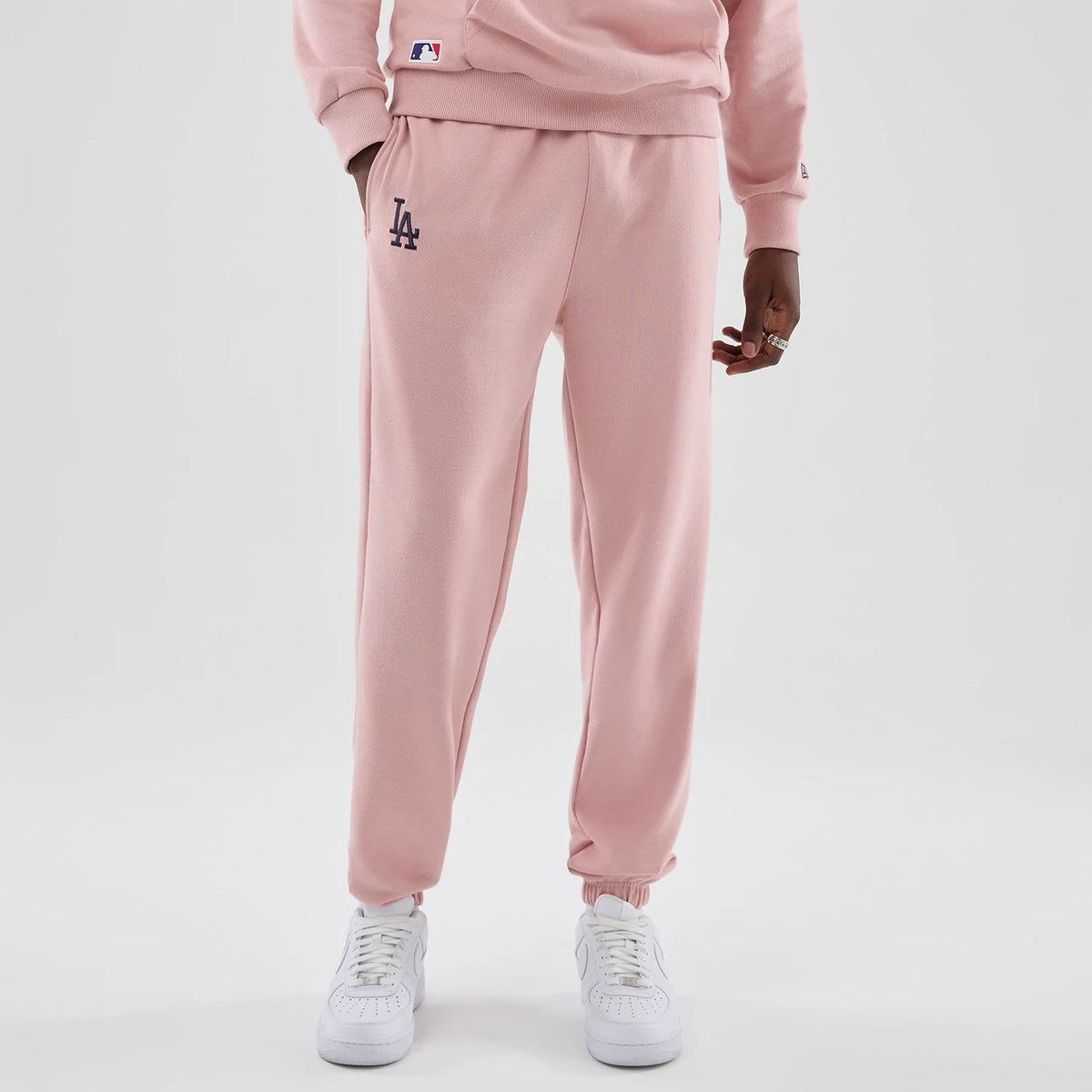 New Era LA Dodgers MLB Midi League Essential Pastel Pink Joggers, front view on a model with LA logo in navy on the left thigh, featuring side pockets, elasticated cuffs, and a matching pastel pink sweatshirt with a small MLB patch near the waist.