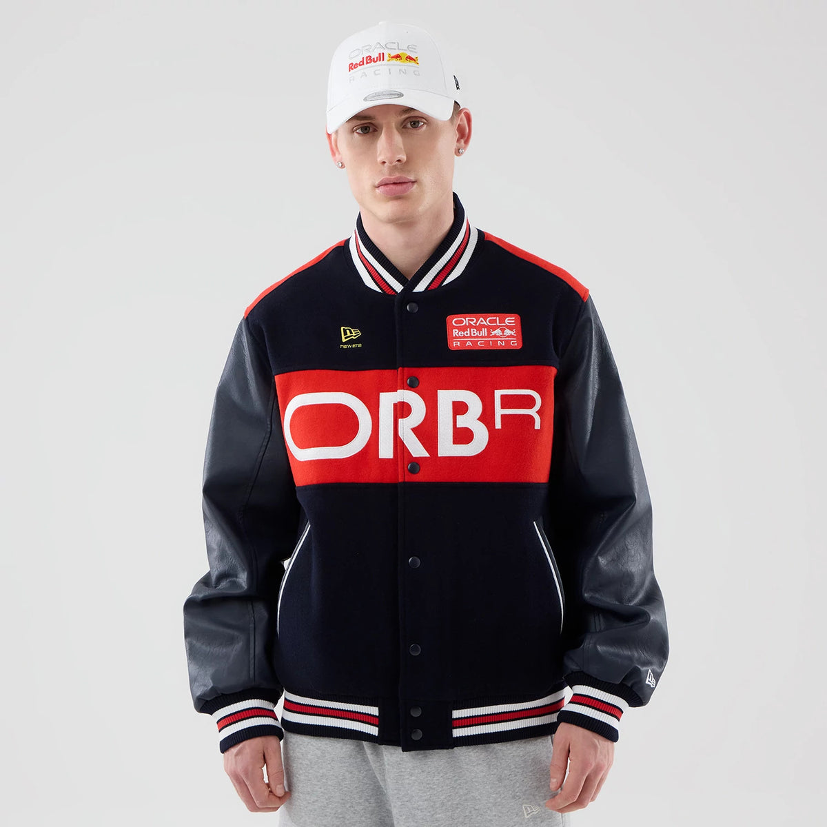 New Era Red Bull Racing wordmark Dark Blue Varsity Jacket, front view on a model showing a large white ORBR print across the chest with a red panel, embroidered New Era logo on the upper chest, and striped ribbing on the cuffs and hem.