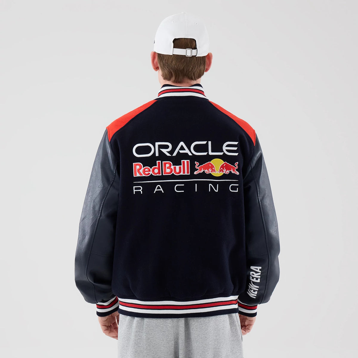 New Era Red Bull Racing wordmark Dark Blue Varsity Jacket, back view on a model with a white cap, showing an embroidered Oracle Red Bull Racing logo centred on the back, red shoulder panels, and New Era lettering on the sleeve cuff.
