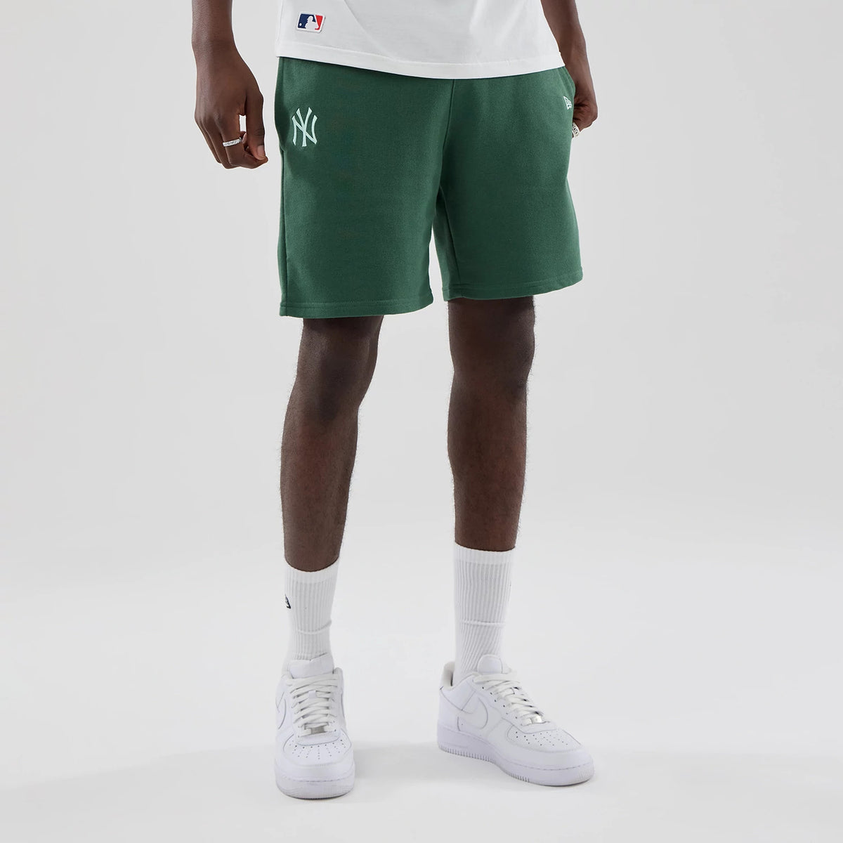 New Era New York Yankees MLB Midi League Essential Dark Green Oversized Shorts, front view on a model highlighting an embroidered white Yankees logo on the left leg, the relaxed fit and mid-length style, paired with white trainers and socks.