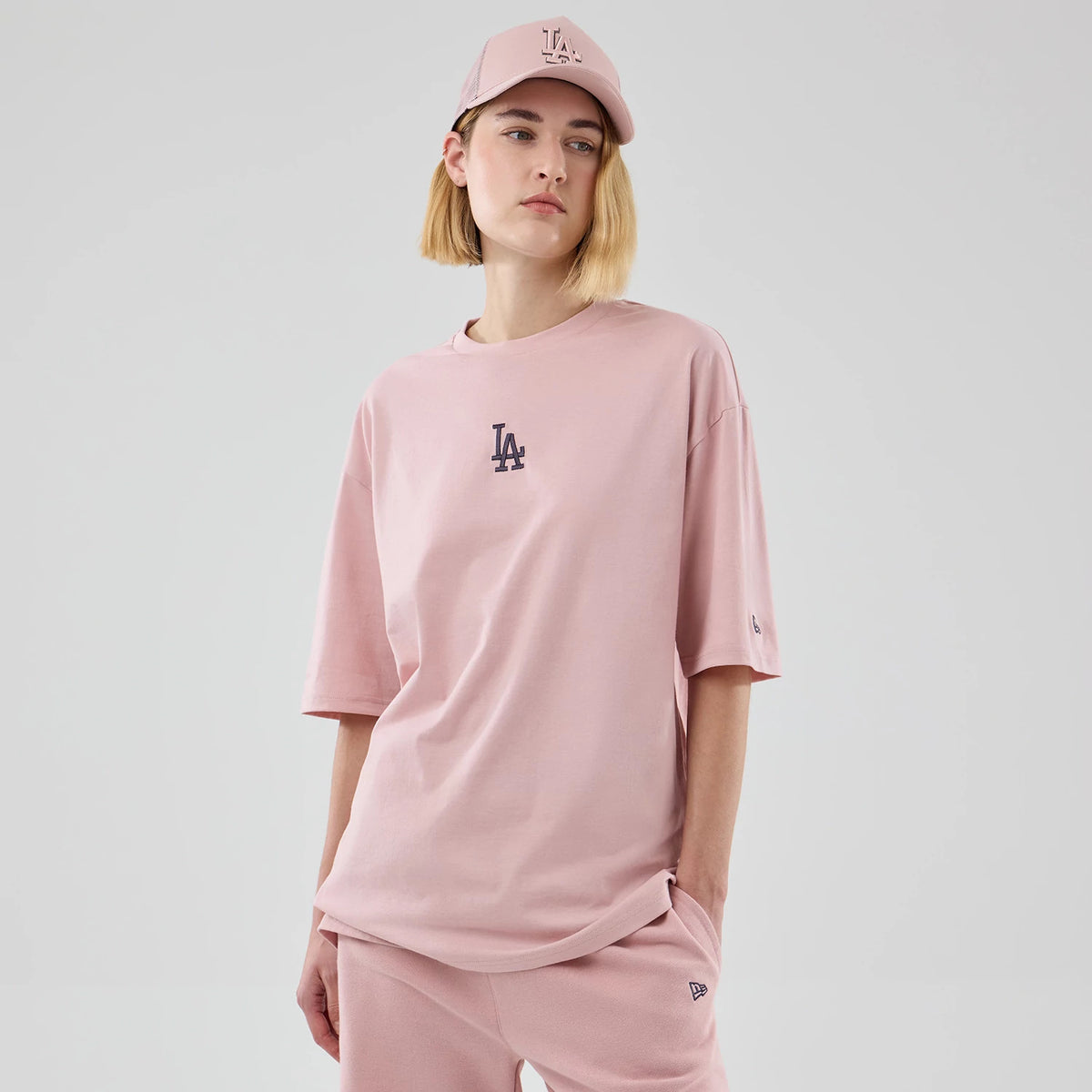 New Era LA Dodgers MLB Midi League Essential Pastel Pink Oversized T-Shirt, front view on a model wearing matching pastel pink trousers and cap, featuring a black embroidered LA logo centred on the chest and a small New Era flag on the left sleeve.