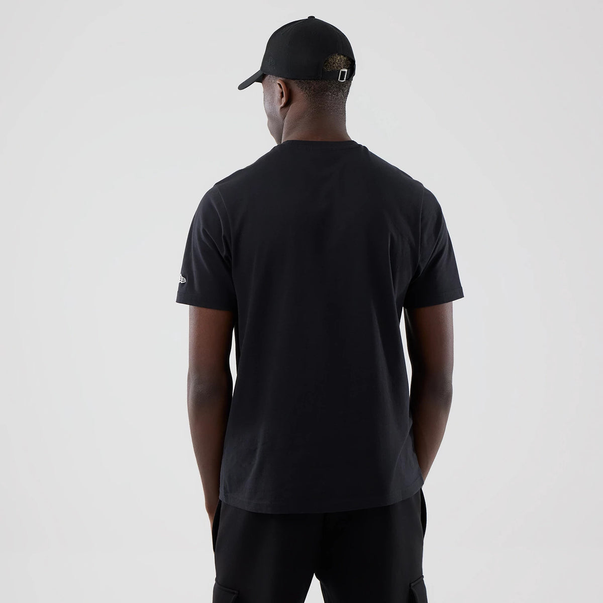 New Era Chicago Bulls NBA Arch Graphic Black T-Shirt, back view on-model showing a plain black rear design with a embroidered New Era logo on the left sleeve, paired with black trousers and a black cap for a coordinated look.