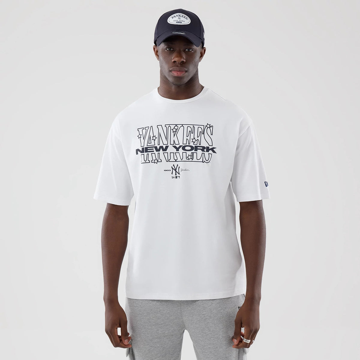 New Era New York Yankees MLB Graphic White Oversized T-Shirt, front view on a model wearing a black cap, showcasing Yankees New York lettering across the chest, a small embroidered New Era logo on the left sleeve, and a monochrome design.