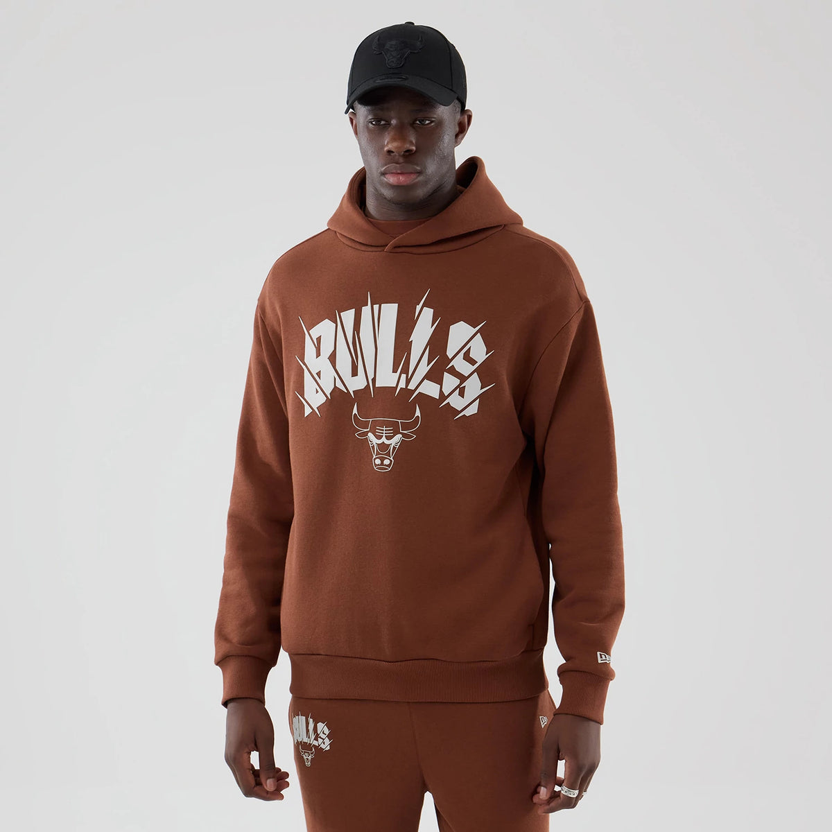 New Era Chicago Bulls NBA Core Plus Brown Pullover Hoodie, front view on a model showing the white Bulls logo with lightning graphics across the chest, a small embroidered New Era logo on the sleeve, and paired with matching brown joggers.